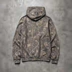A hooded sweatshirt laid flat on a textured gray concrete surface, shown from the back. The hoodie features an all-over camouflage pattern in earthy brown, gray, beige, and muted olive tones.