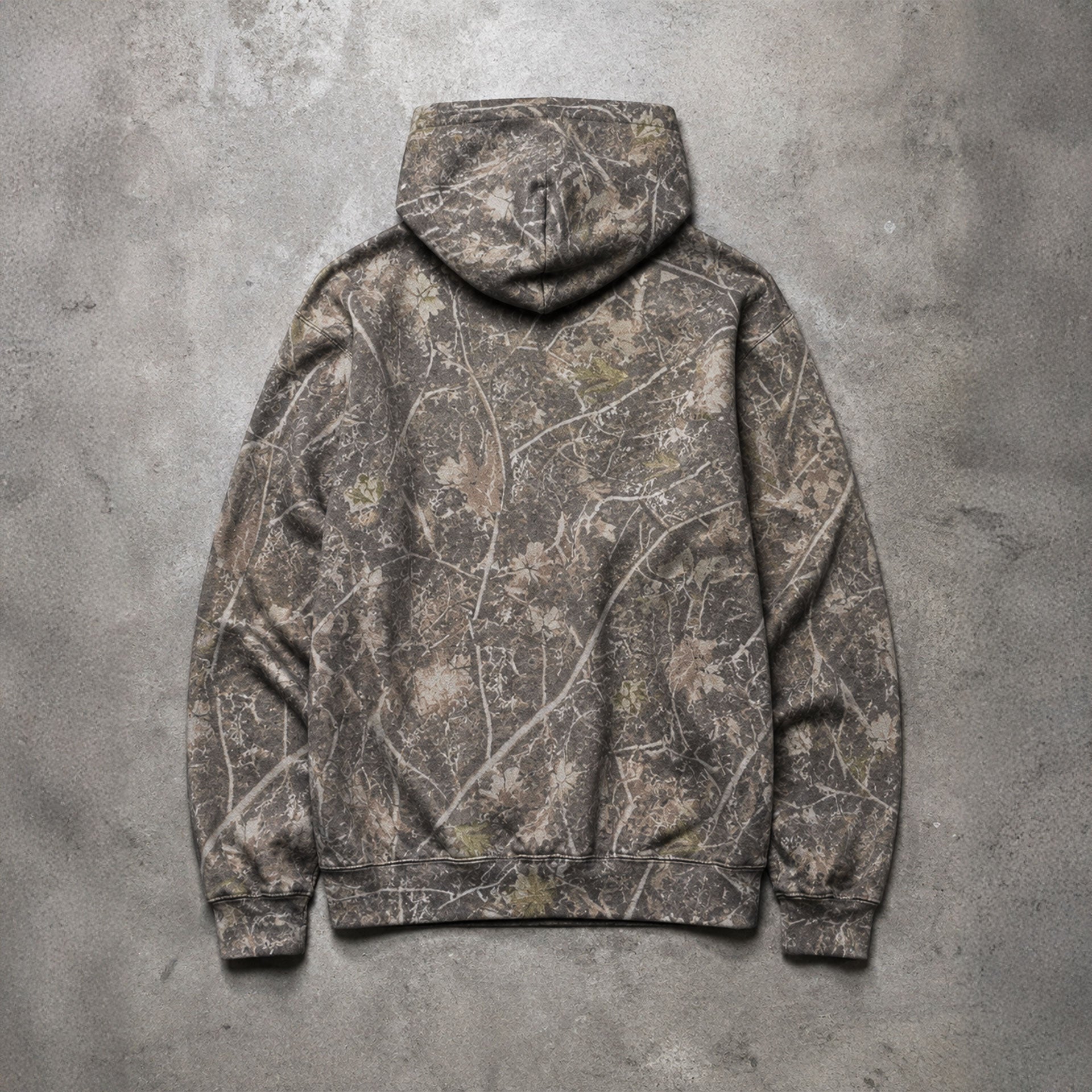 A hooded sweatshirt laid flat on a textured gray concrete surface, shown from the back. The hoodie features an all-over camouflage pattern in earthy brown, gray, beige, and muted olive tones.