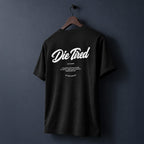 A black short-sleeve t-shirt hanging on a wooden hanger against a dark navy blue wall, featuring a white graphic print on the back that reads 'Die Tired' with smaller text below.