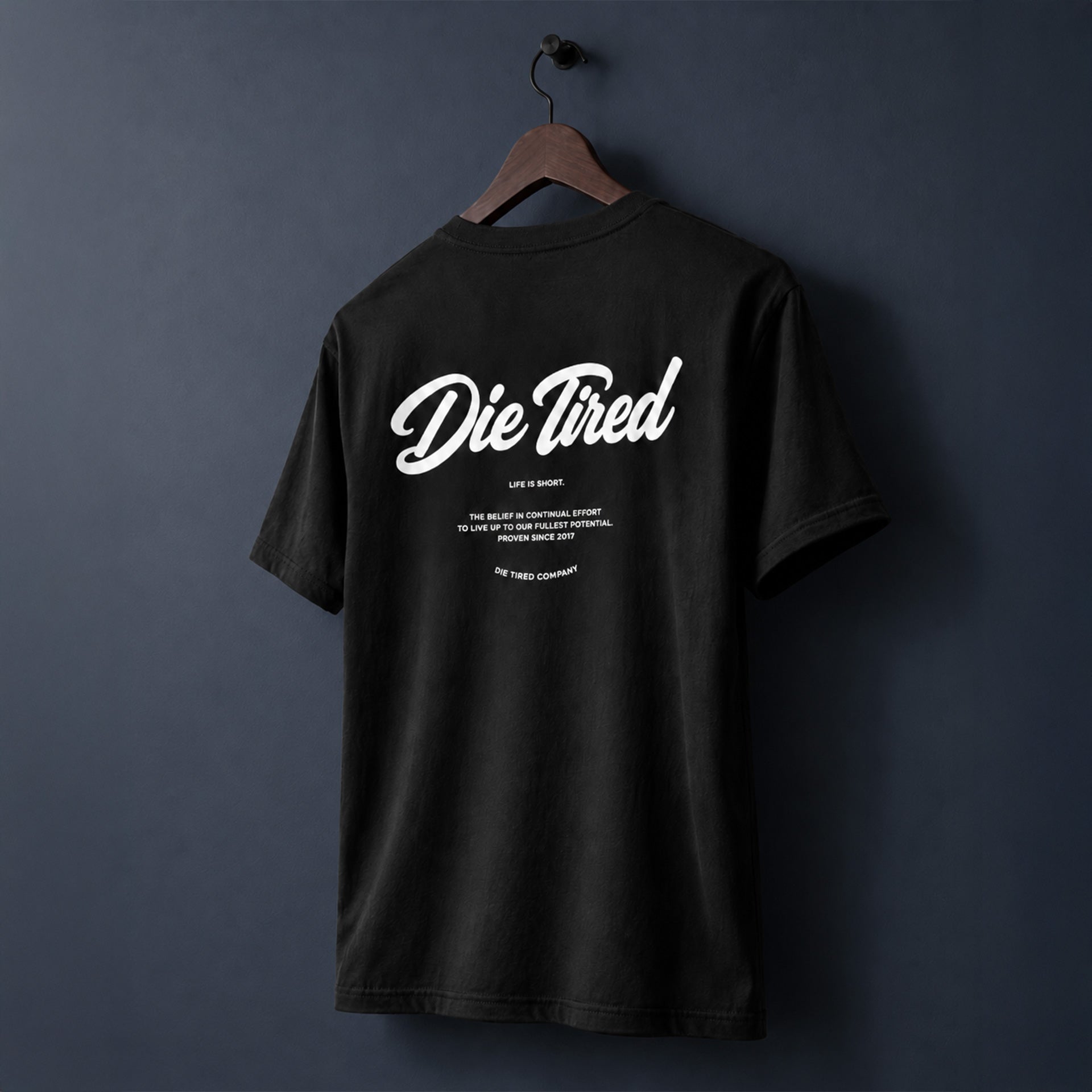 A black short-sleeve t-shirt hanging on a wooden hanger against a dark navy blue wall, featuring a white graphic print on the back that reads 'Die Tired' with smaller text below.
