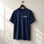 A navy blue short-sleeve t-shirt hanging on a wooden hanger against a light beige wall with soft window shadow patterns. The shirt features a small white printed  logo on the left chest that reads 'Die Tired EST. 2017'.