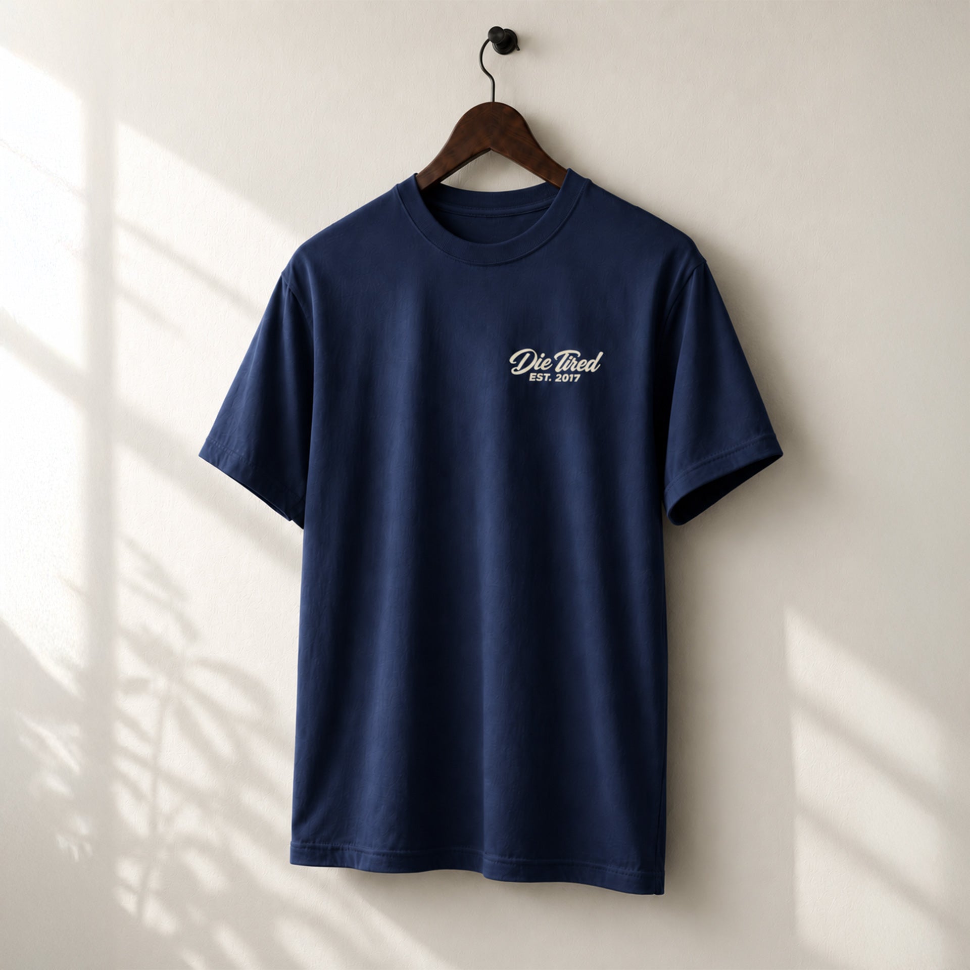A navy blue short-sleeve t-shirt hanging on a wooden hanger against a light beige wall with soft window shadow patterns. The shirt features a small white printed  logo on the left chest that reads 'Die Tired EST. 2017'.
