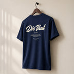 A navy blue t-shirt hanging on a wooden hanger against a warm beige wall, featuring a white graphic print on the back that reads 'Die Tired' with smaller text below.