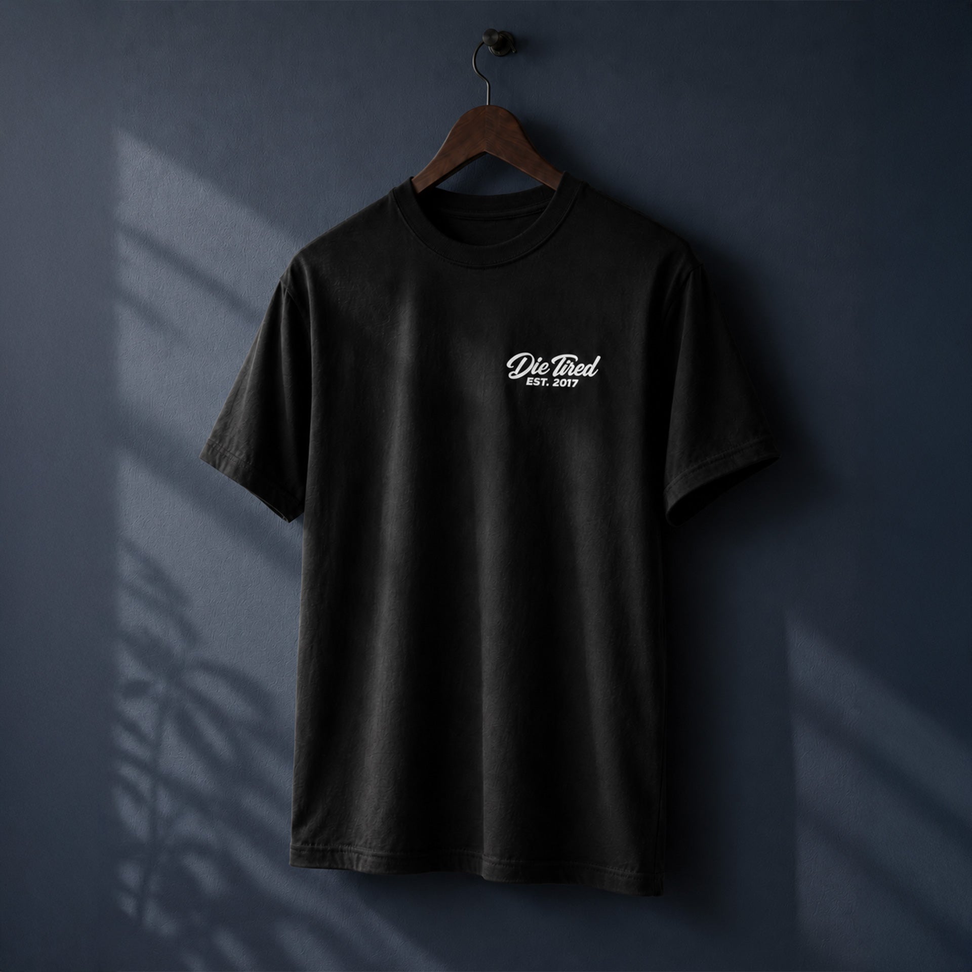A black short-sleeve t-shirt hanging on a wooden hanger against a dark blue wall, featuring a small white 'DIE TIRED EST. 2017" hand written cursive logo printed on the chest.