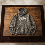 A dark gray camouflage hoodie with a white 'Die Tired' graphic on the chest, laid flat on a rustic wooden board over a dark patterned rug.