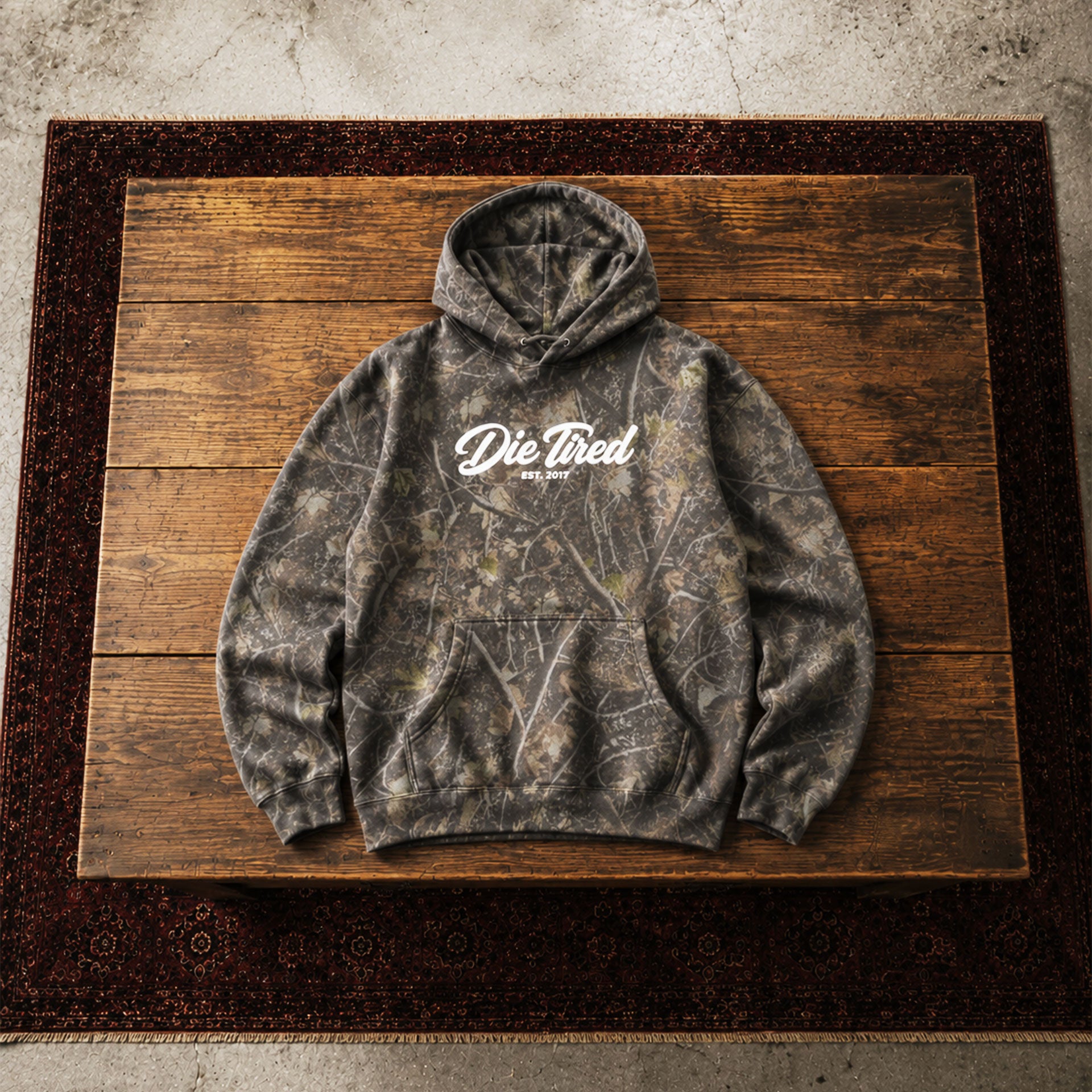 A dark gray camouflage hoodie with a white 'Die Tired' graphic on the chest, laid flat on a rustic wooden board over a dark patterned rug.