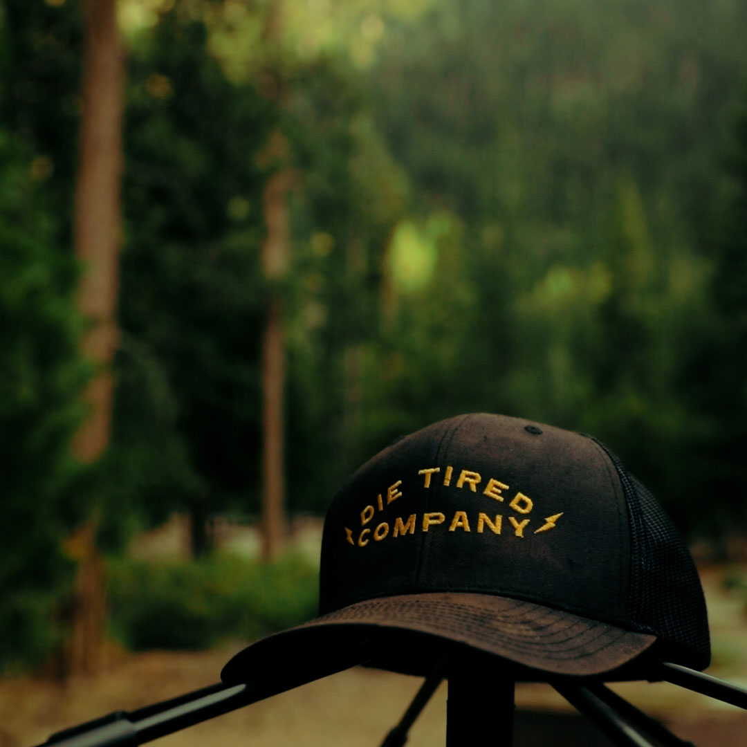 A black trucker-style cap with yellow embroidered text reading 'DIE TIRED COMPANY' and a small lightning bolt, photographed outdoors against a blurred forest background.