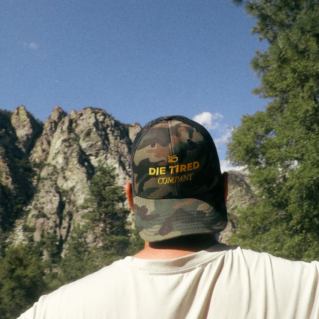 A person viewed from behind wearing a camouflage trucker cap with yellow embroidered text reading 'DIE TIRED COMPANY' against a mountain and tree landscape.