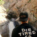 A man viewed from behind wearing a black baseball cap and a black t-shirt with 'DIE TIRED COMPANY' printed on the back, standing in front of a rocky cliff with a waterfall.
