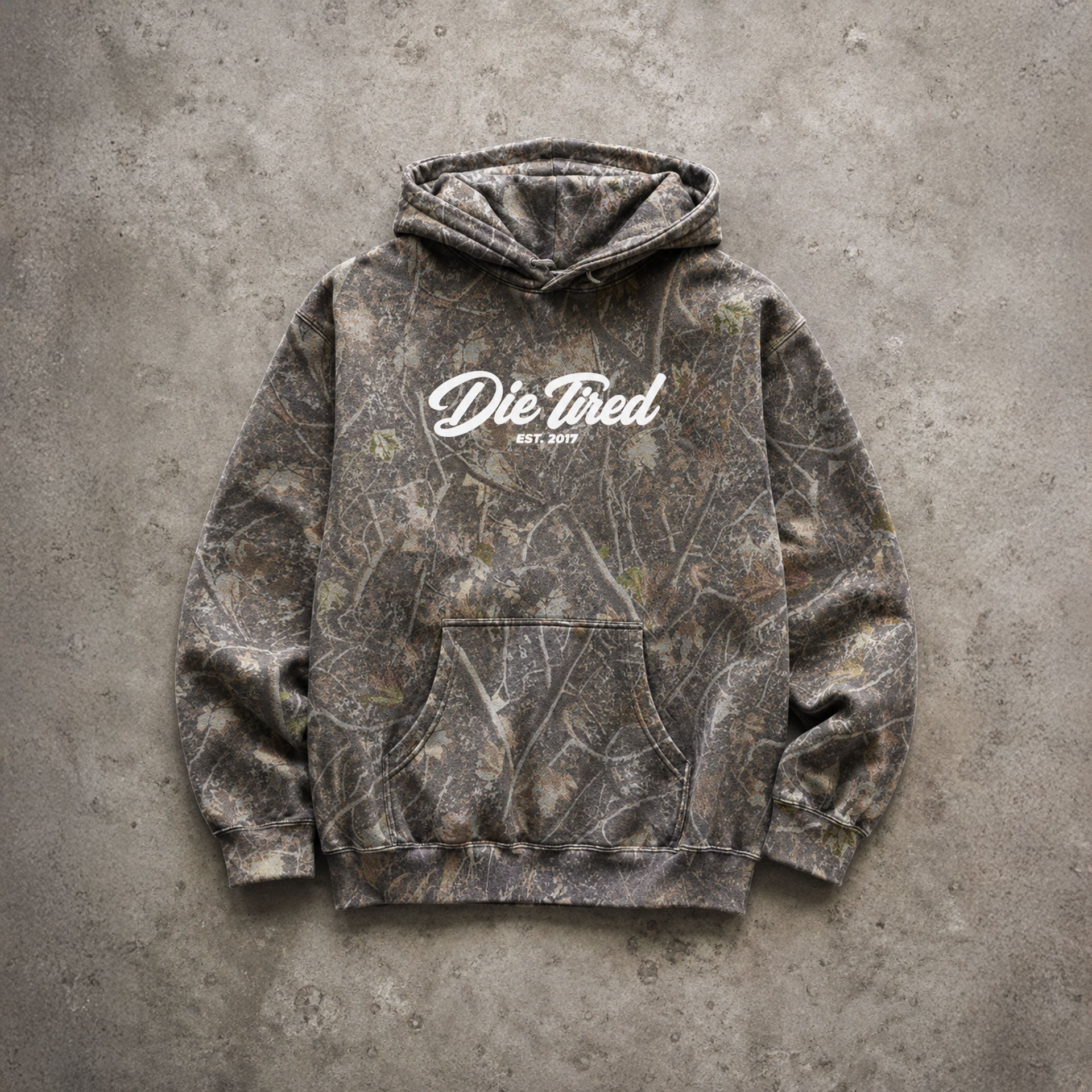 A pullover hoodie with a brown and gray tree-branch camouflage pattern, featuring a white 'Die Thied ESTD 2019' logo printed on the chest, laid flat on a textured gray concrete surface.