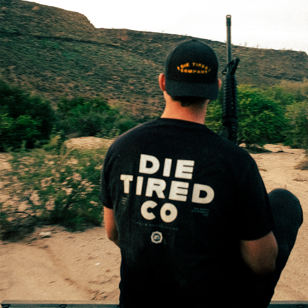 Person wearing a black t-shirt with 'DIE TIRED CO' text, holding a rifle in a desert landscape.