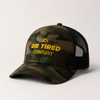 Camouflage trucker hat with 'Die Tired Company' text on a light gray background