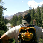 A person seen from behind wearing a camouflage trucker hat with yellow 'DIE TIRED COMPANY' text and a yellow and black hiking backpack, looking out at a mountain river valley surrounded by pine trees.