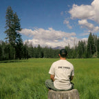 Man sitting on a tree stump in a beautiful pine forest meadow scene with clear blue skies with puffy white clouds. Die Tired graphic tee with screen printed logo on back that reads "Die Tired".
