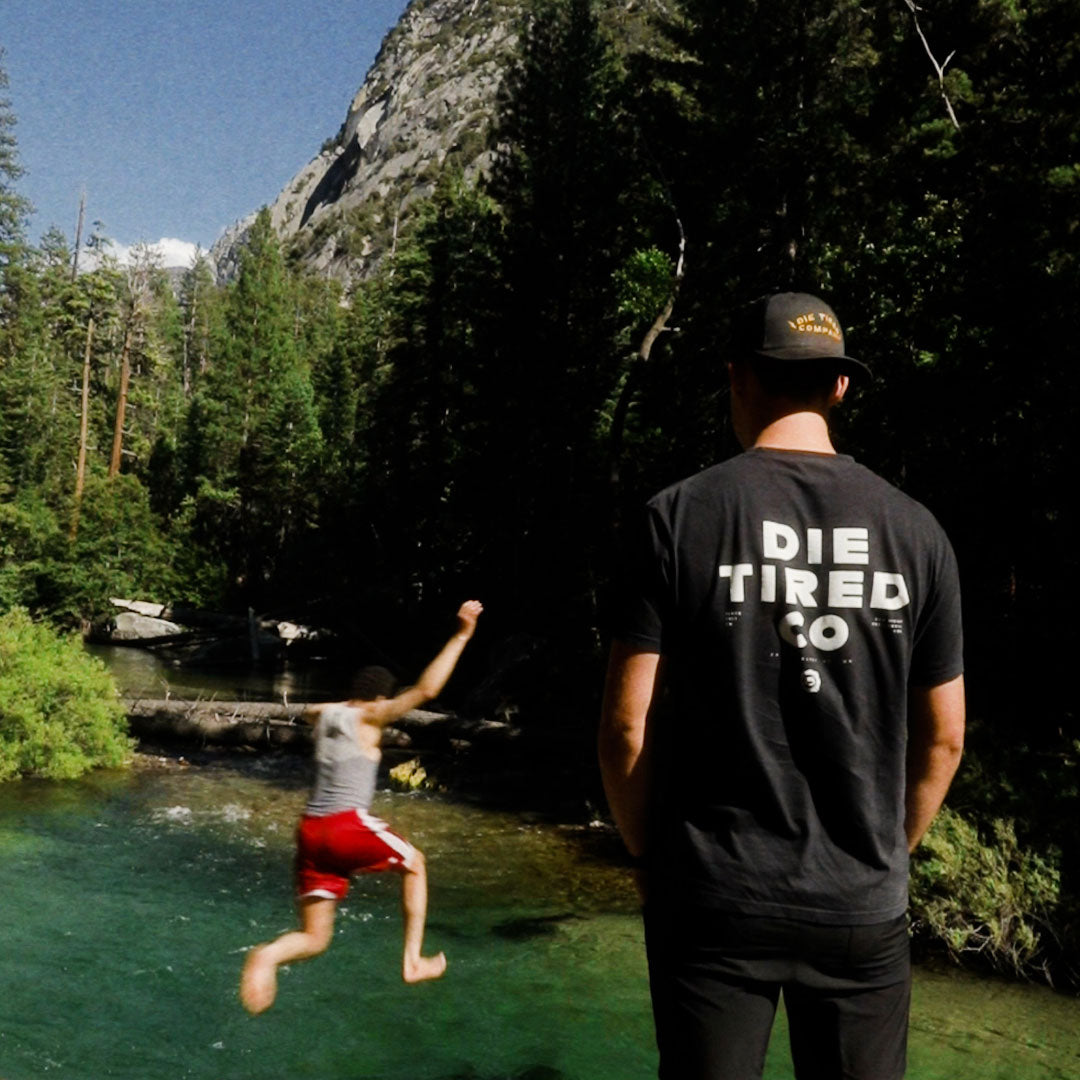 A man stands with his back to the camera wearing a black t-shirt with 'DIE TIRED CO' printed in white, overlooking a person jumping into a clear green river surrounded by a dense forest and rocky mountain.