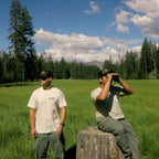 two friends surveying beautiful scenic meadow with pine forest slightly blurred in the background. Both are wearing Die Tired Co natural white graphic tee shirts with Die Tired Company trucker hats.