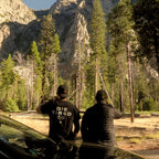 Two men standing outdoors in front of a mountain and pine trees, viewed from behind. One man wears a black T-shirt with the text 'DIE TIRED CO' on the back, and the other wears a black puffer jacket.