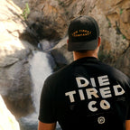 A man viewed from behind wearing a black trucker cap and a black t-shirt with 'DIE TIRED COMPANY' printed in gold and white on the back, set against a natural rocky waterfall background.