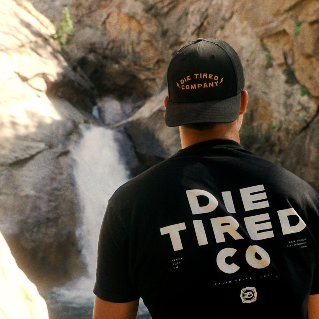 A man viewed from behind wearing a black trucker cap and a black t-shirt with 'DIE TIRED COMPANY' printed in gold and white on the back, set against a natural rocky waterfall background.